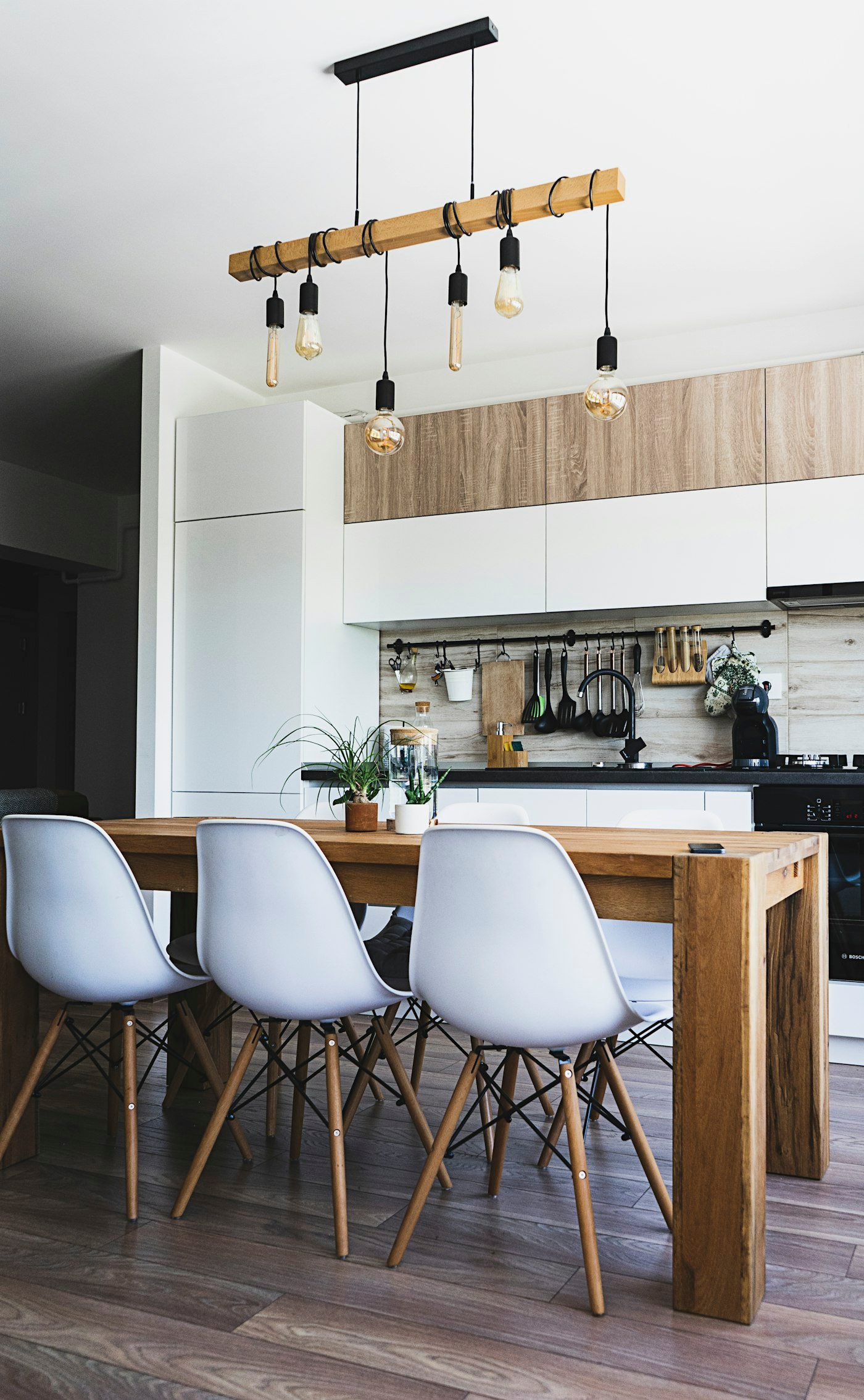 A wood dining table with white chairs, and a kitchen of white cupboards and wood panels